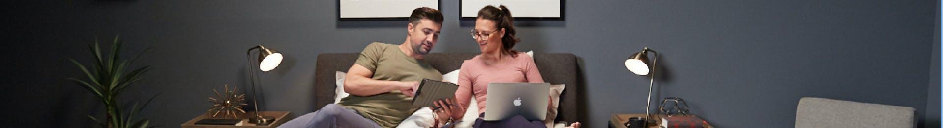 Smiling couple sitting on a Sleeping Duck Mattress in a contemporary bedroom. One person is reading a touch pad tablet, while the other uses a laptop.